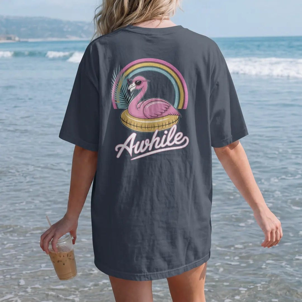 Woman on the beach in a Midnight tee, back view: a pink flamingo in sunglasses lounging on a pool float, with "STAY AWHILE" in neon-tube script, printed in faded, cracked cream, mustard and turquoise with a neon pink accent over a faded rainbow arc.
