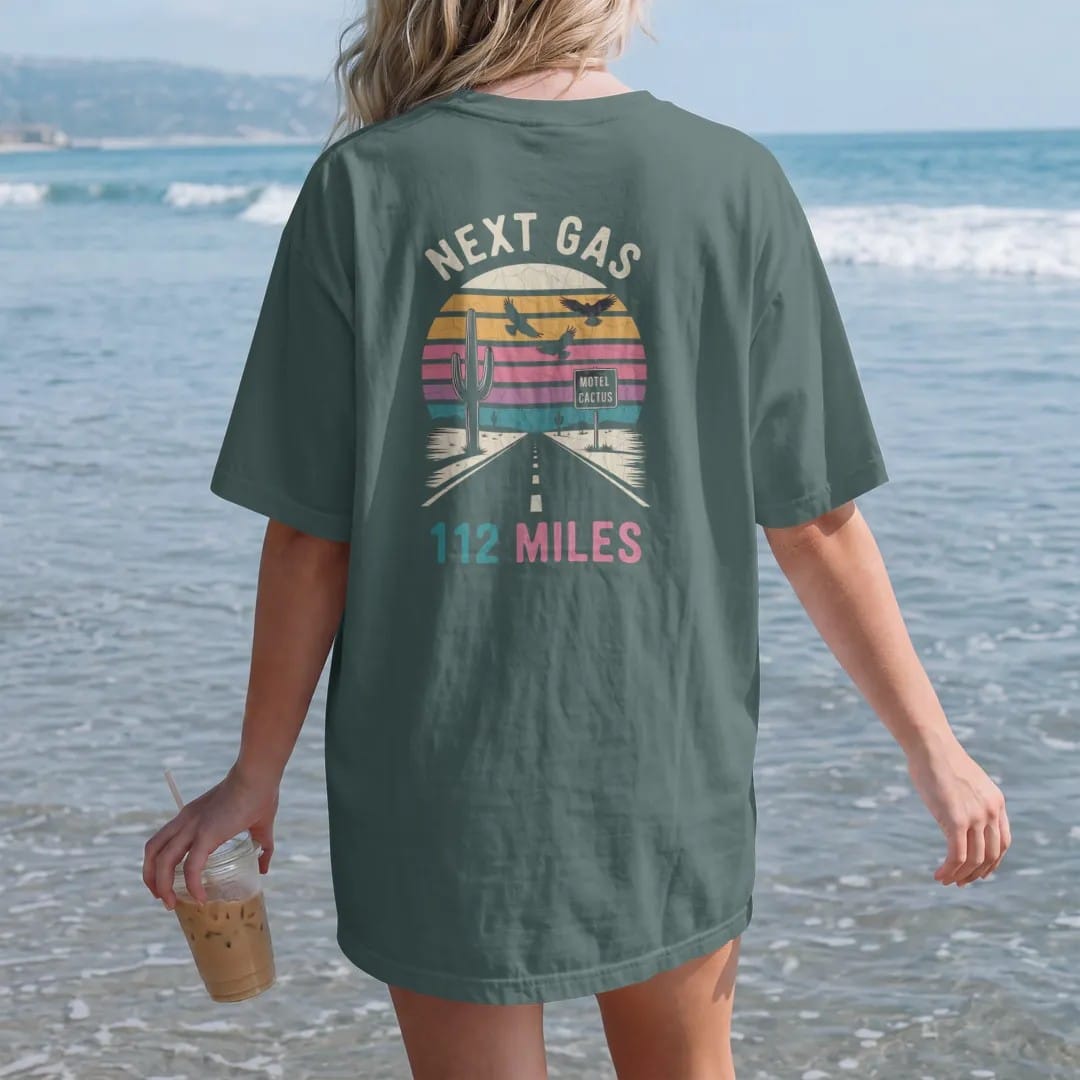 Woman on the beach in a Blue Spruce tee, back view: a desert highway sign beside saguaro cactus under a sunset, with "NEXT GAS" and "112 MILES" in chunky 70s lettering and small 70s caps, printed in faded, cracked cream, mustard and turquoise with…