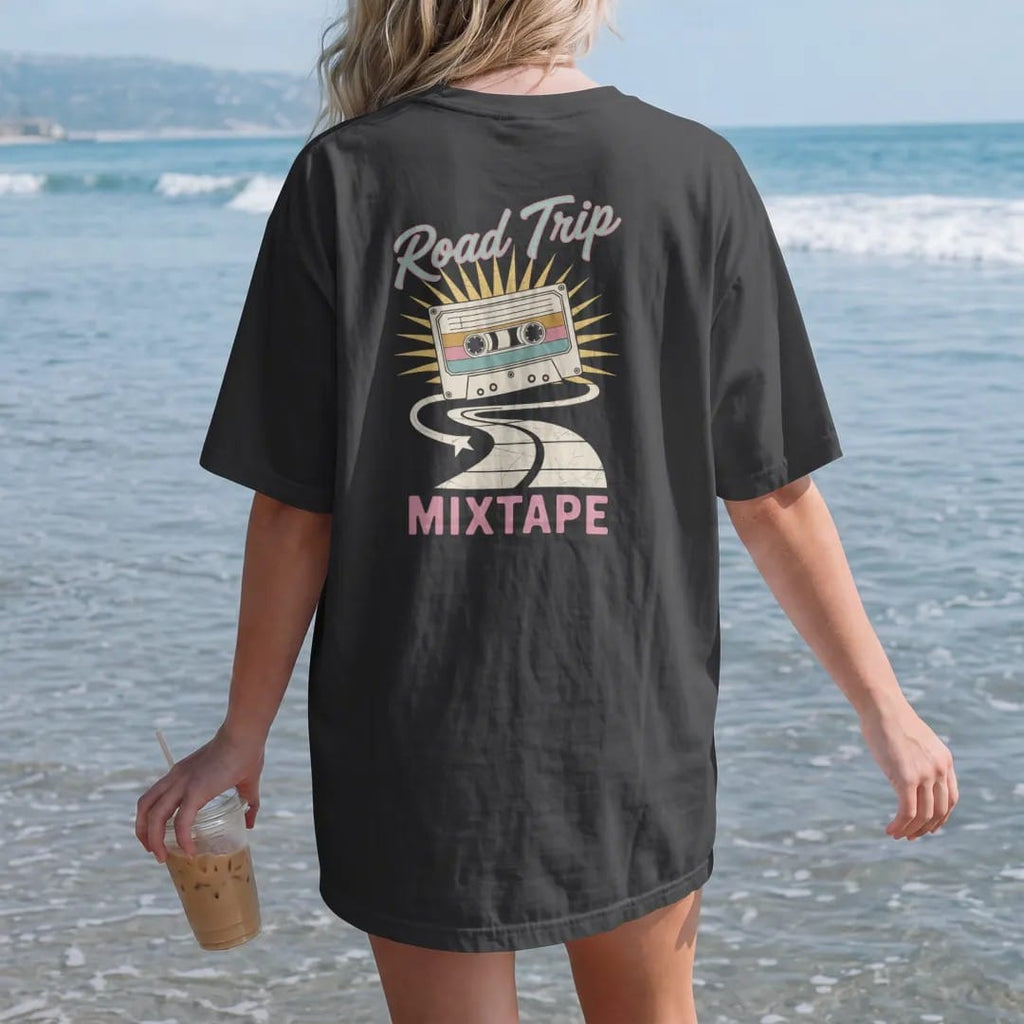 Woman on the beach in a Black tee, back view: a cassette tape whose ribbon unspools into a winding road, with "ROAD TRIP" and "MIXTAPE" in neon-tube script and small 70s caps, printed in faded, cracked cream, mustard and turquoise with a neon pink…