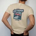 Man wearing a Sandstone tee, seen from the back: a rustic motor-lodge sign over mountains in fall color, with "LEAF PEEPER" and "LODGE" in chunky 70s lettering and small 70s caps, printed in faded, cracked navy, rust red and dusty teal with a neon…