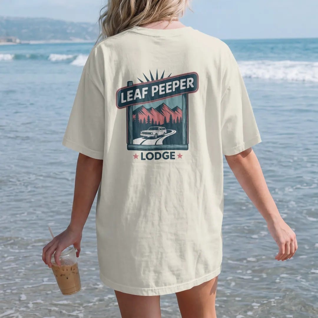 Woman on the beach in a Ivory tee, back view: a rustic motor-lodge sign over mountains in fall color, with "LEAF PEEPER" and "LODGE" in chunky 70s lettering and small 70s caps, printed in faded, cracked navy, rust red and dusty teal with a neon…
