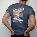 Man wearing a Navy tee, seen from the back: a snowy motel with lit windows and a glowing neon sign, with "SNOWED IN" and "MOTOR LODGE" in neon-tube script and small 70s caps, printed in faded, cracked cream, mustard and turquoise with a neon pink…