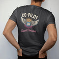 Man wearing a Black tee, seen from the back: a winged pilot badge with crossed chips bag and paper map, with "CO-PILOT" and "SNACK DIVISION" in chunky 70s lettering and brush script, printed in faded, cracked cream, mustard and turquoise with a…