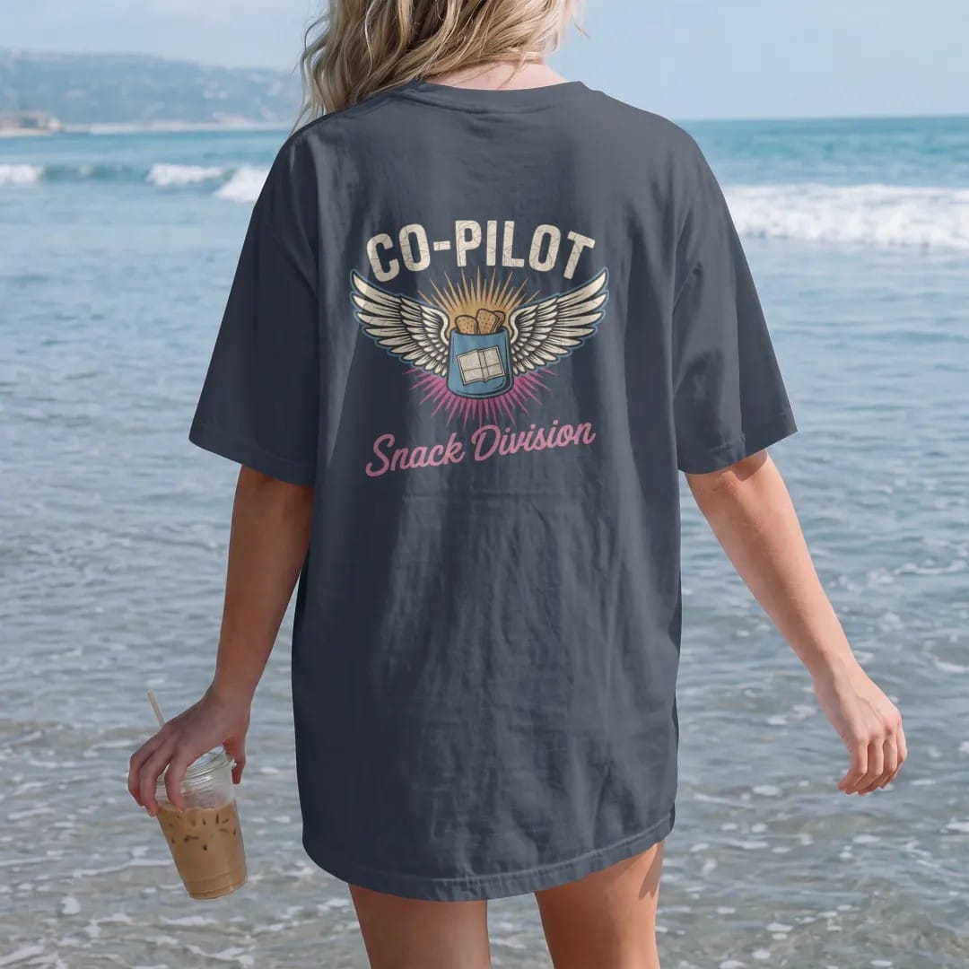 Woman on the beach in a Midnight tee, back view: a winged pilot badge with crossed chips bag and paper map, with "CO-PILOT" and "SNACK DIVISION" in chunky 70s lettering and brush script, printed in faded, cracked cream, mustard and turquoise with a…