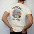 Man wearing a Ivory tee, seen from the back: a fanned stack of vintage motel postcards held by a rubber band, with "POSTCARD" and "COLLECTOR" in chunky 70s lettering and small 70s caps, printed in faded, cracked navy, rust red and dusty teal with a…