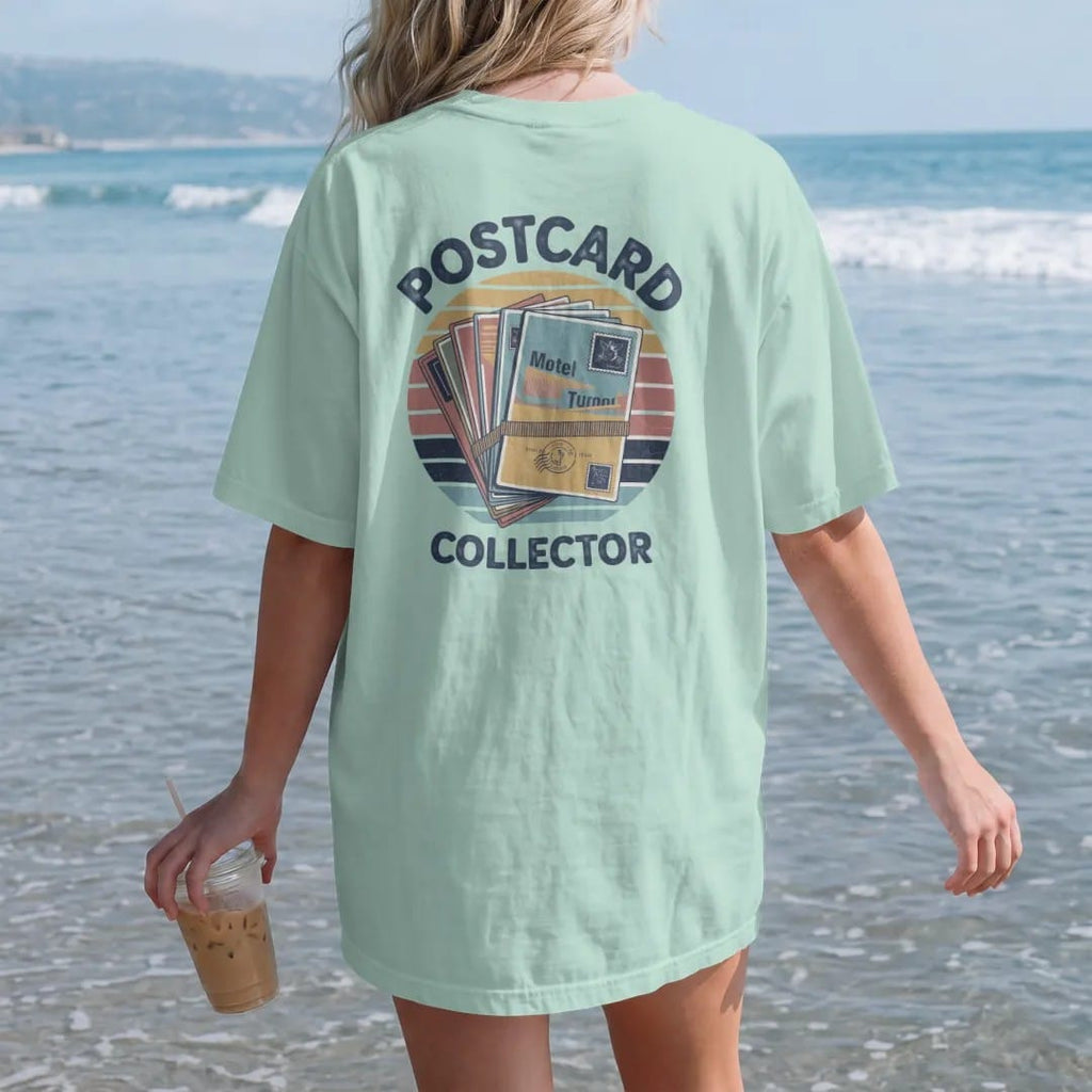 Woman on the beach in a Chalky Mint tee, back view: a fanned stack of vintage motel postcards held by a rubber band, with "POSTCARD" and "COLLECTOR" in chunky 70s lettering and small 70s caps, printed in faded, cracked navy, rust red and dusty teal…
