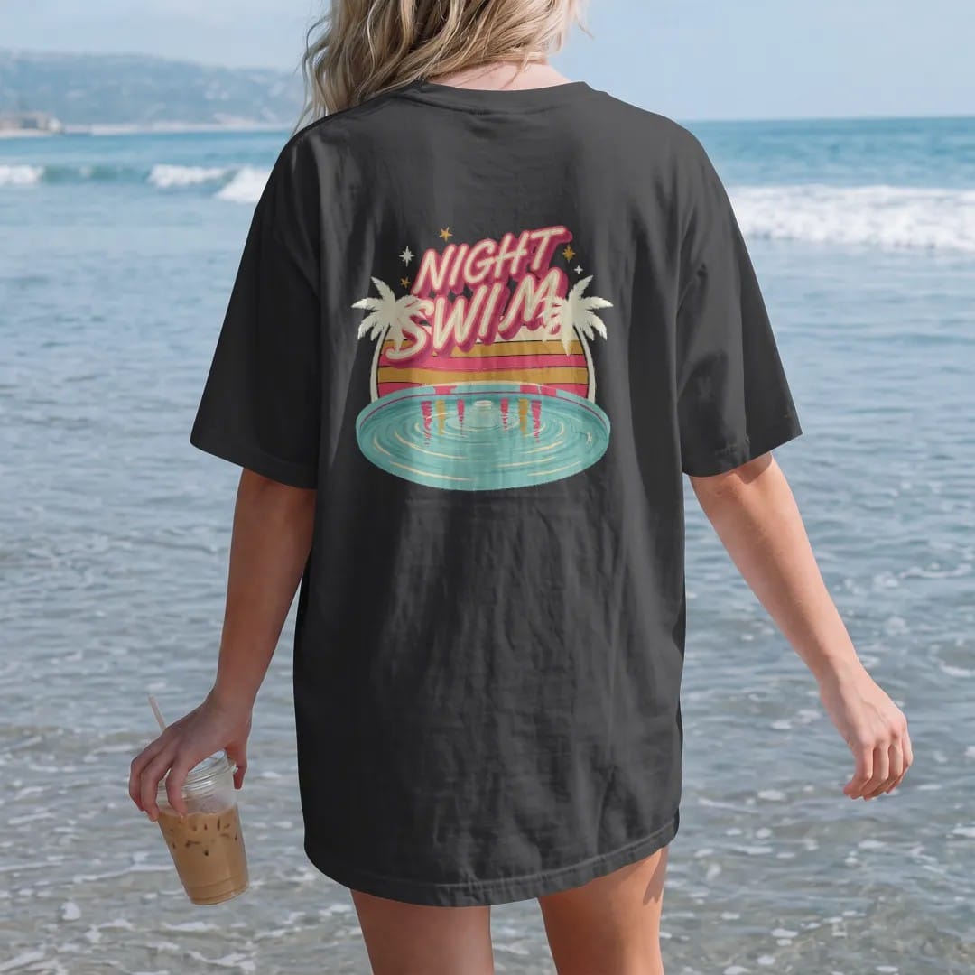 Woman on the beach in a Black tee, back view: a glowing motel pool at night reflecting a neon sign, with "NIGHT SWIM" in neon-tube script, printed in faded, cracked cream, mustard and turquoise with a neon pink accent over faded sunset stripes.