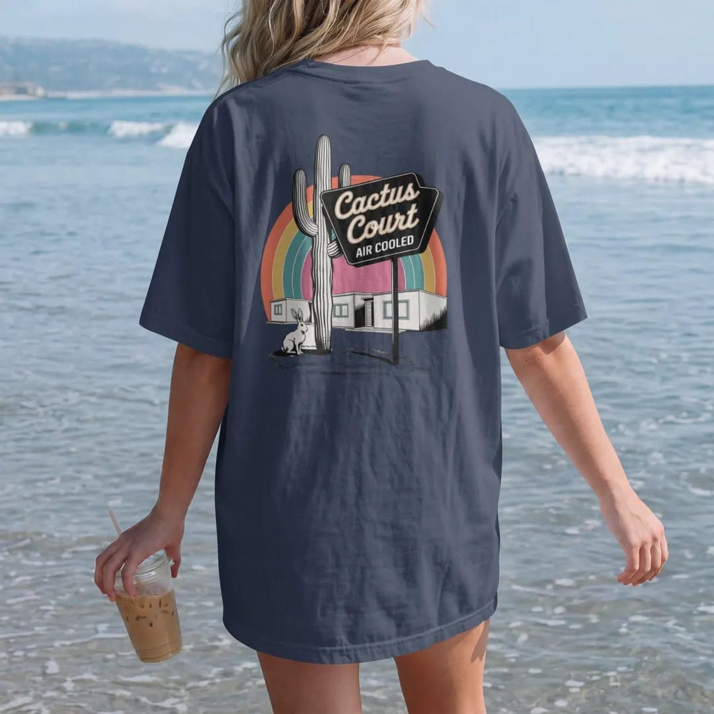 Woman on the beach in a Navy tee, back view: a saguaro-shaped neon motel sign above adobe bungalows, with "CACTUS COURT" and "AIR COOLED" in neon-tube script and small 70s caps, printed in faded, cracked cream, mustard and turquoise with a neon…