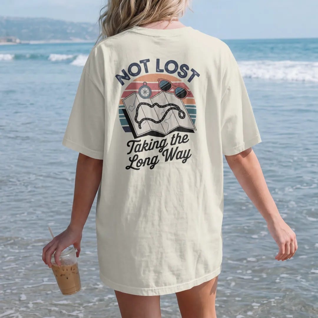 Woman on the beach in a Ivory tee, back view: an unfolded road map with a wobbly looping route, with "NOT LOST" and "TAKING THE LONG WAY" in chunky 70s lettering and brush script, printed in faded, cracked navy, rust red and dusty teal with a neon…