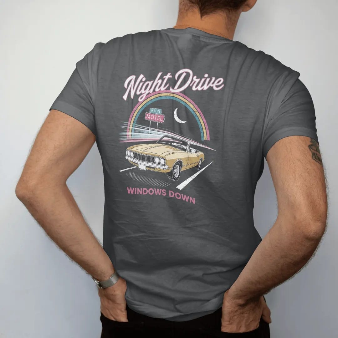 Man wearing a Graphite tee, seen from the back: a convertible on a dark highway with neon streaks, with "NIGHT DRIVE" and "WINDOWS DOWN" in neon-tube script and small 70s caps, printed in faded, cracked cream, mustard and turquoise with a neon pink…