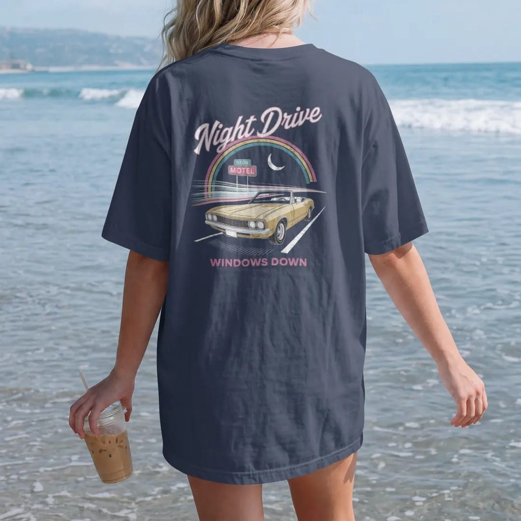 Woman on the beach in a Navy tee, back view: a convertible on a dark highway with neon streaks, with "NIGHT DRIVE" and "WINDOWS DOWN" in neon-tube script and small 70s caps, printed in faded, cracked cream, mustard and turquoise with a neon pink…