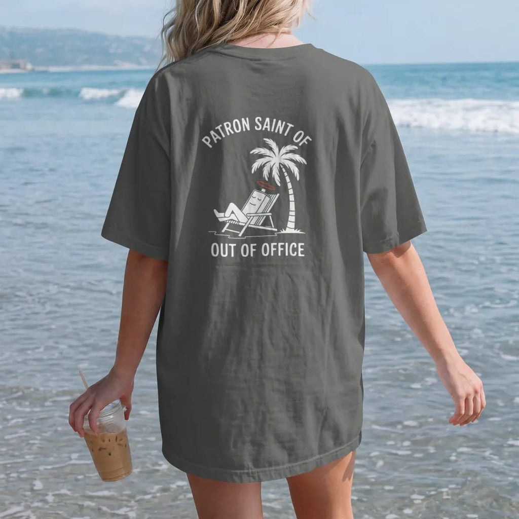 Woman on the beach in a Pepper tee, back view: minimal single-line drawing of a sleepy battery with a tiny halo lounging in a beach chair under a single palm tree, with "PATRON SAINT OF / OUT OF OFFICE" in small spaced-out caps, printed in bone…