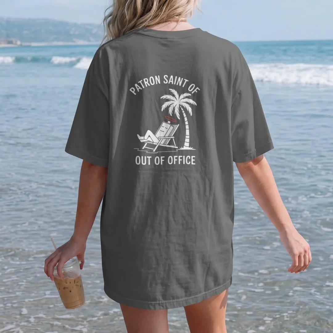 Woman on the beach in a Pepper tee, back view: minimal single-line drawing of a sleepy battery with a tiny halo lounging in a beach chair under a single palm tree, with "PATRON SAINT OF / OUT OF OFFICE" in small spaced-out caps, printed in bone…