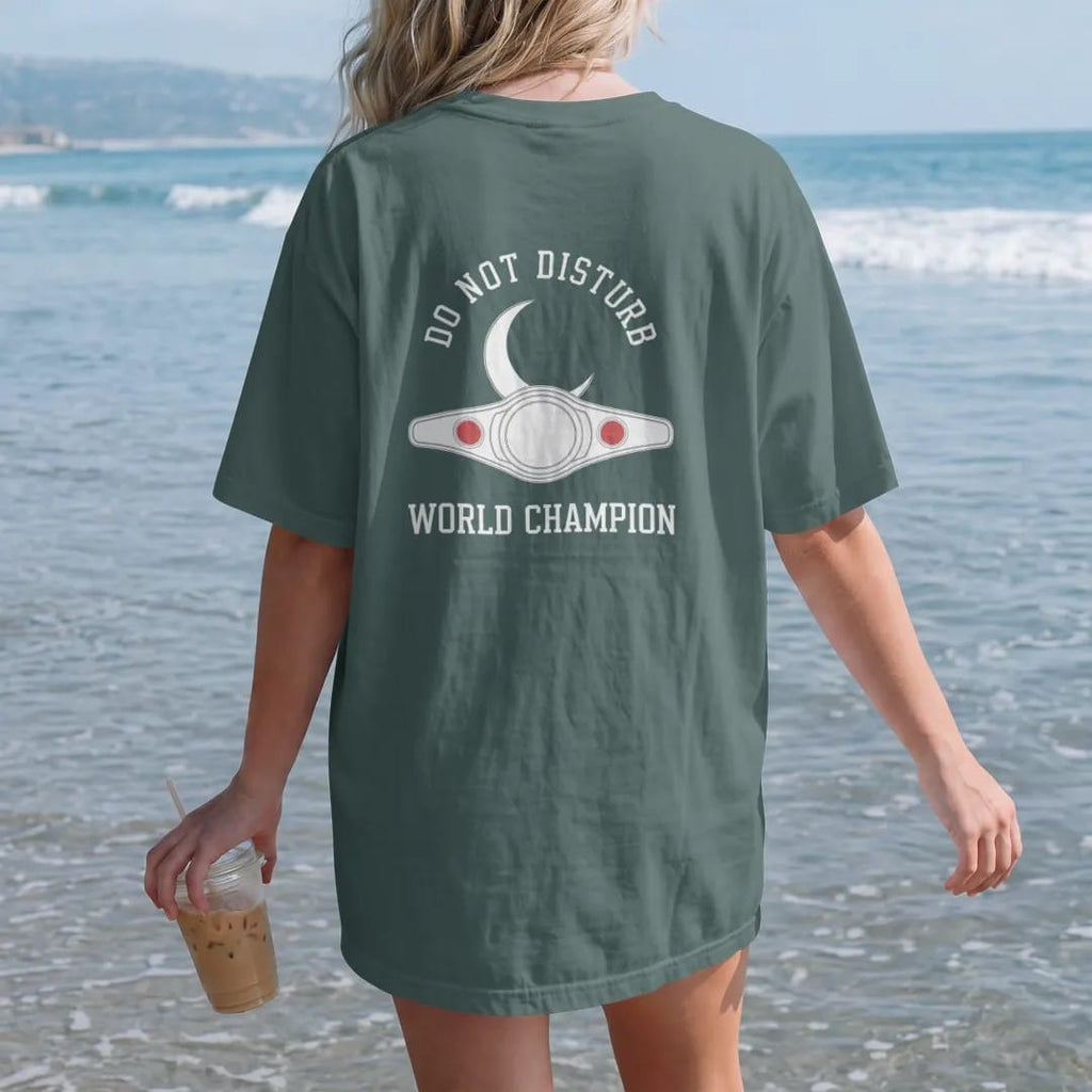 Woman on the beach in a Blue Spruce tee, back view: minimal single-line drawing of a crescent moon resting on a slim outline championship belt, with "DO NOT DISTURB / WORLD CHAMPION" in small spaced-out caps, printed in bone white with a…