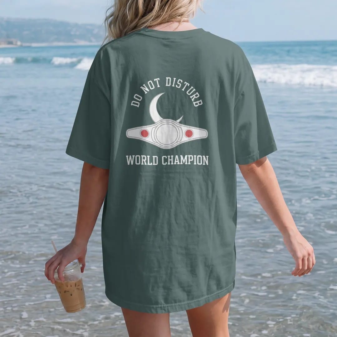 Woman on the beach in a Blue Spruce tee, back view: minimal single-line drawing of a crescent moon resting on a slim outline championship belt, with "DO NOT DISTURB / WORLD CHAMPION" in small spaced-out caps, printed in bone white with a…