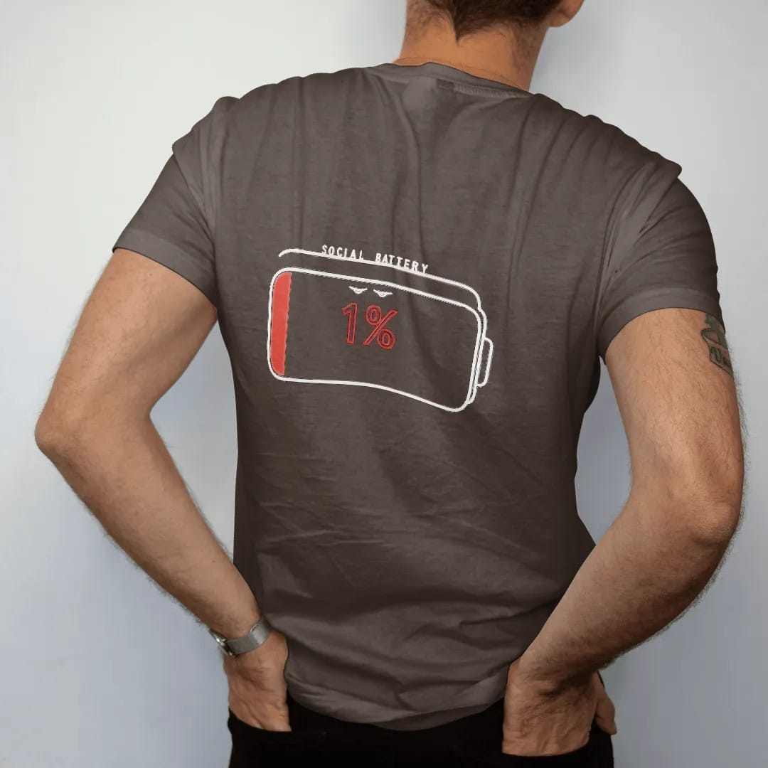 Man wearing a Espresso tee, seen from the back: minimal single-line drawing of a large outline battery icon with droopy eyes and one thin sliver of charge, with "SOCIAL BATTERY / 1%" in small spaced-out caps, printed in bone white with a…