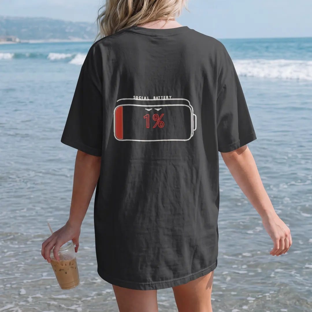Woman on the beach in a Black tee, back view: minimal single-line drawing of a large outline battery icon with droopy eyes and one thin sliver of charge, with "SOCIAL BATTERY / 1%" in small spaced-out caps, printed in bone white with a low-battery…