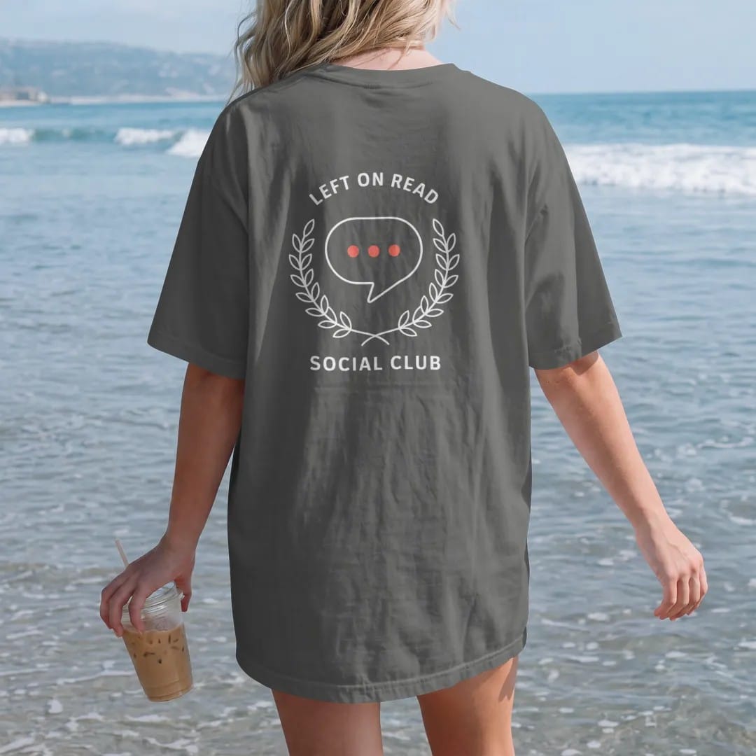 Woman on the beach in a Pepper tee, back view: minimal single-line drawing of a single chat bubble with three typing dots inside a thin laurel wreath, with "LEFT ON READ / SOCIAL CLUB" in small spaced-out caps, printed in bone white with a…