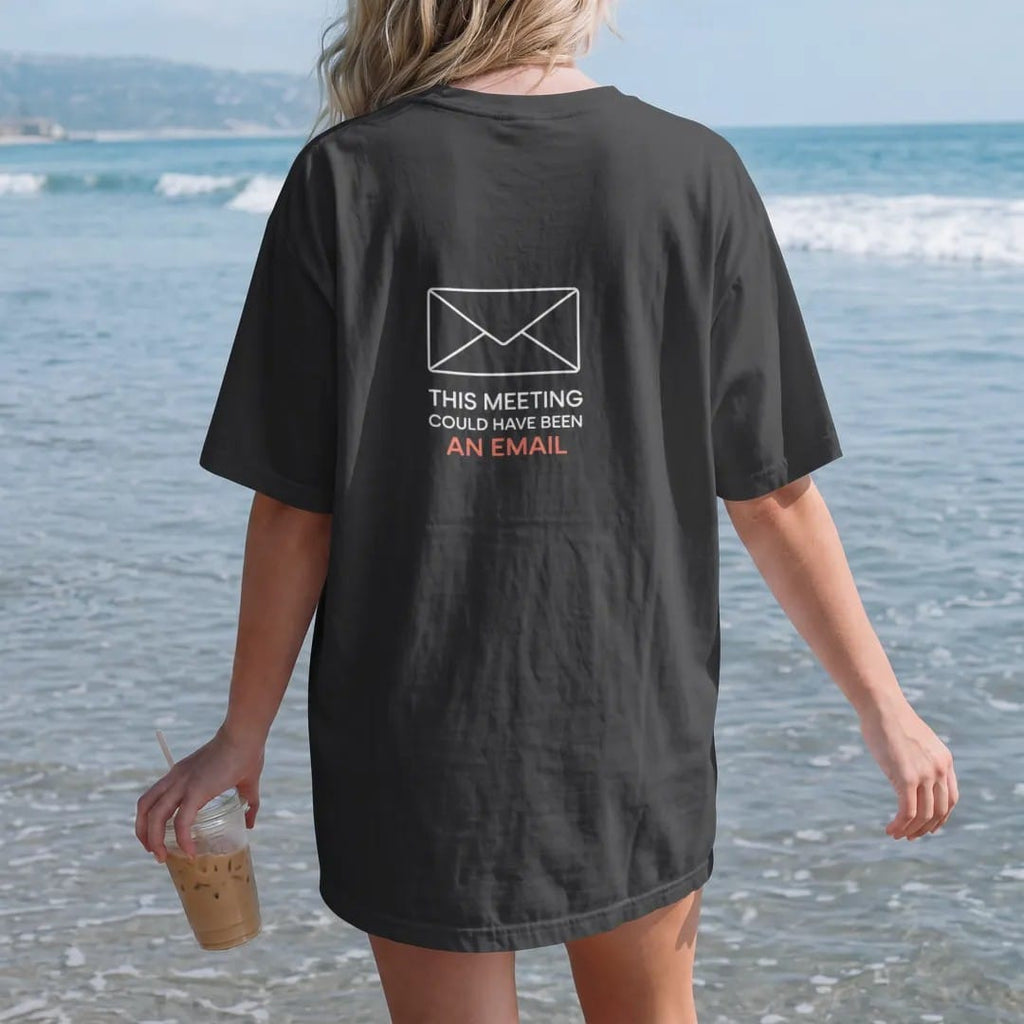 Woman on the beach in a Black tee, back view: minimal single-line drawing of a tiny envelope outline, with "THIS MEETING / COULD HAVE BEEN / AN EMAIL" in small spaced-out caps, printed in bone white with a low-battery red accent on the envelope.