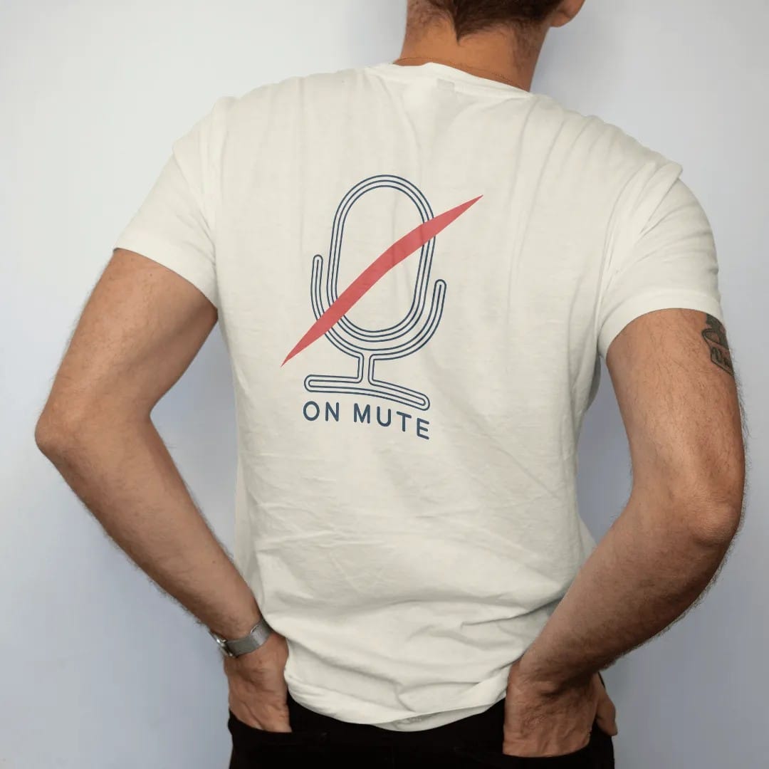 Man wearing a Ivory tee, seen from the back: minimal single-line drawing of a microphone outline with one diagonal slash through it, with "YOU'RE ON MUTE" in small spaced-out caps, printed in ink navy with a battery red accent on the slash.