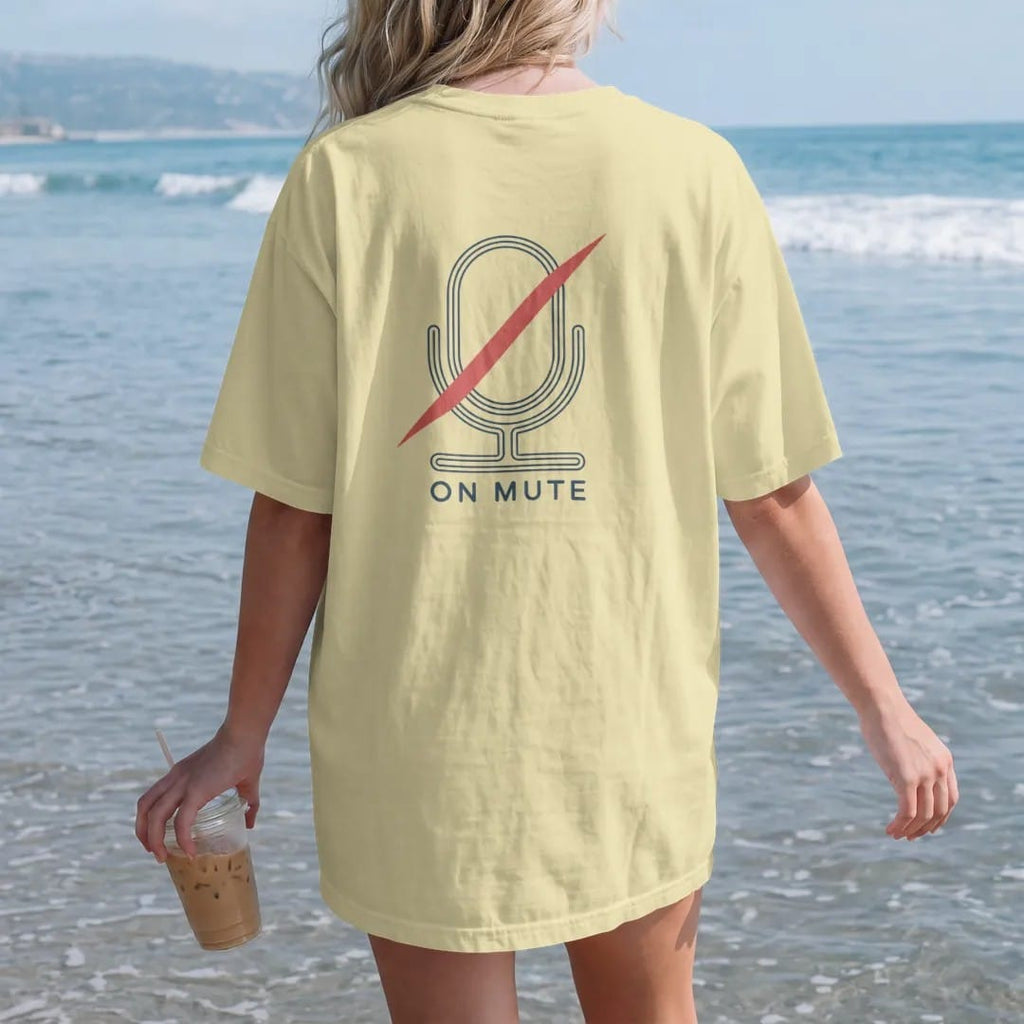 Woman on the beach in a Butter tee, back view: minimal single-line drawing of a microphone outline with one diagonal slash through it, with "YOU'RE ON MUTE" in small spaced-out caps, printed in ink navy with a battery red accent on the slash.