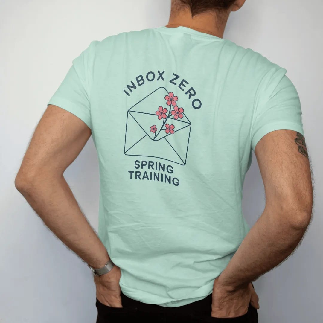 Man wearing a Chalky Mint tee, seen from the back: minimal single-line drawing of an envelope outline with a small cherry blossom sprig tucked on top, with "INBOX ZERO / SPRING TRAINING" in small spaced-out caps, printed in ink navy with a battery…