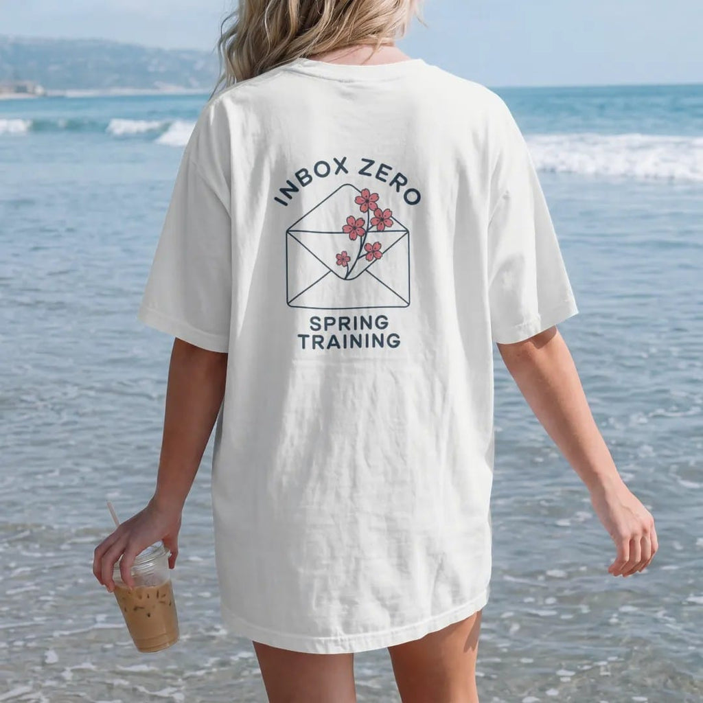 Woman on the beach in a White tee, back view: minimal single-line drawing of an envelope outline with a small cherry blossom sprig tucked on top, with "INBOX ZERO / SPRING TRAINING" in small spaced-out caps, printed in ink navy with a battery red…
