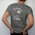 Man wearing a Pepper tee, seen from the back: minimal single-line drawing of a closed laptop half buried in sand under a single palm tree and a small sun, with "OUT OF OFFICE / ALL SUMMER" in small spaced-out caps, printed in bone white with a…