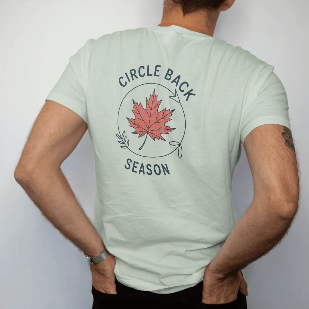 Man wearing a Bay tee, seen from the back: minimal single-line drawing of a single autumn leaf riding a thin looping circular arrow, with "CIRCLE BACK / SEASON" in small spaced-out caps, printed in ink navy with a battery red accent on the leaf.