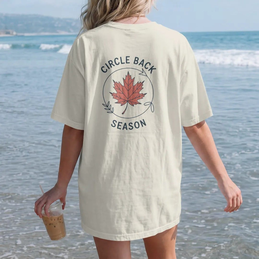 Woman on the beach in a Ivory tee, back view: minimal single-line drawing of a single autumn leaf riding a thin looping circular arrow, with "CIRCLE BACK / SEASON" in small spaced-out caps, printed in ink navy with a battery red accent on the leaf.