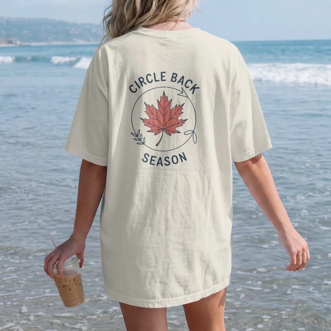 Woman on the beach in a Ivory tee, back view: minimal single-line drawing of a single autumn leaf riding a thin looping circular arrow, with "CIRCLE BACK / SEASON" in small spaced-out caps, printed in ink navy with a battery red accent on the leaf.