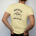 Man wearing a Butter tee, seen from the back: minimal single-line drawing of two coffee mugs clinking with a tiny heart between them, with "WORK BESTIE / SINCE DAY ONE" in small spaced-out caps, printed in ink navy with a battery red accent on the…
