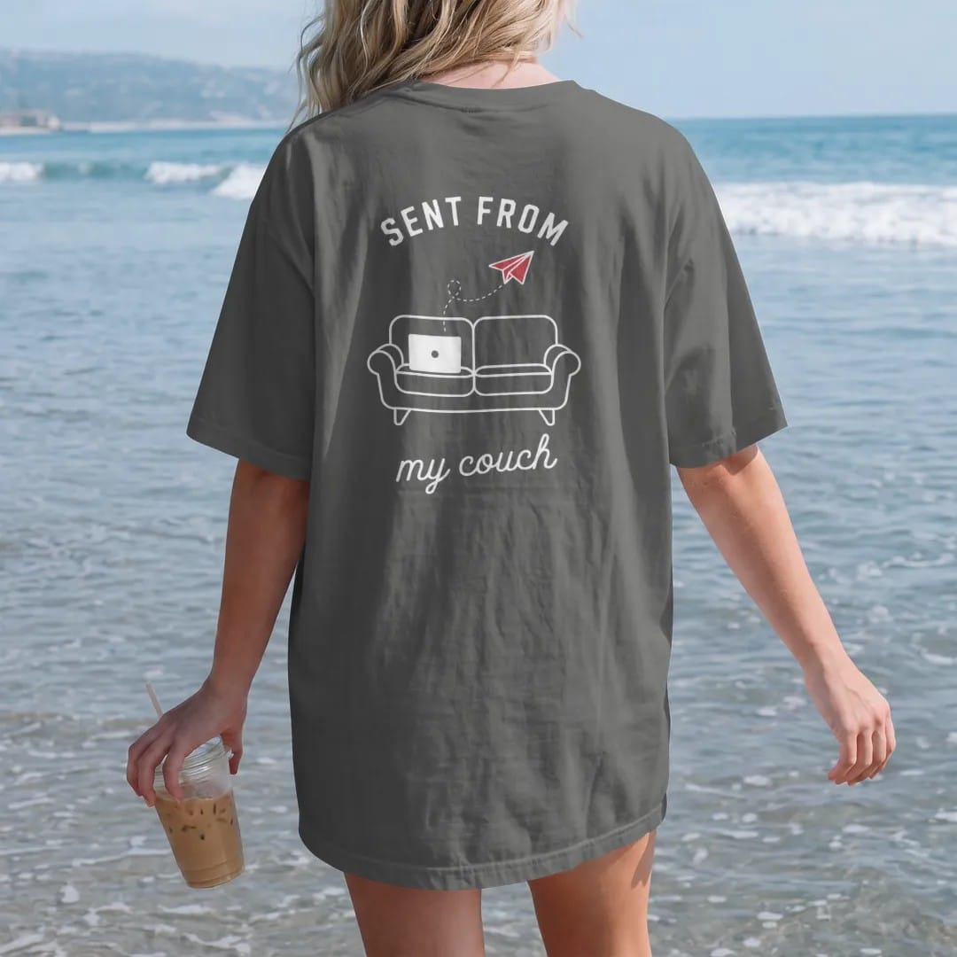 Woman on the beach in a Pepper tee, back view: minimal single-line drawing of a simple sofa outline with a laptop and a paper plane floating up from it, with "SENT FROM / MY COUCH" in small spaced-out caps, printed in bone white with a low-battery…