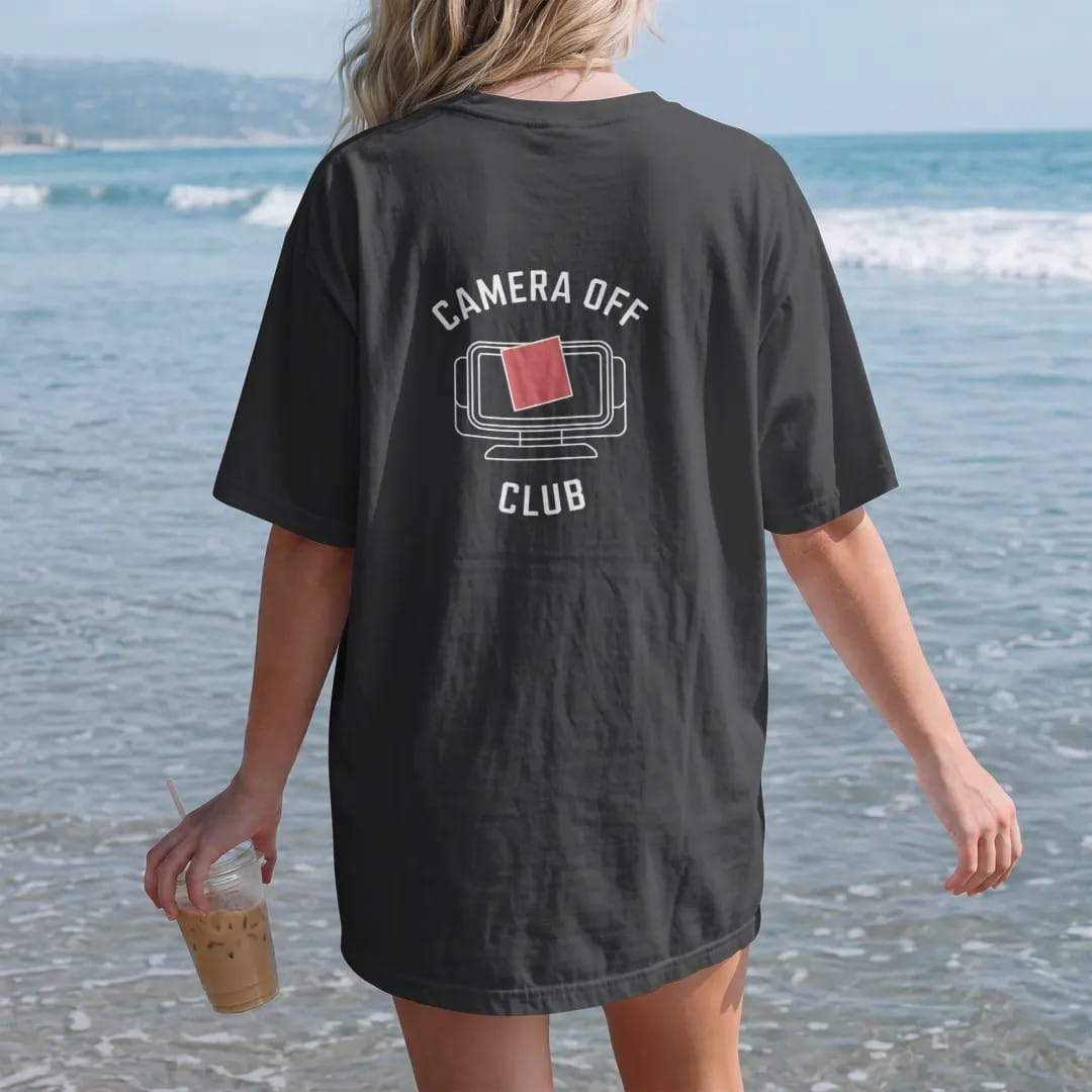 Woman on the beach in a Black tee, back view: minimal single-line drawing of a small webcam outline covered by a square sticky note, with "CAMERA OFF / CLUB" in small spaced-out caps, printed in bone white with a low-battery red accent on the…