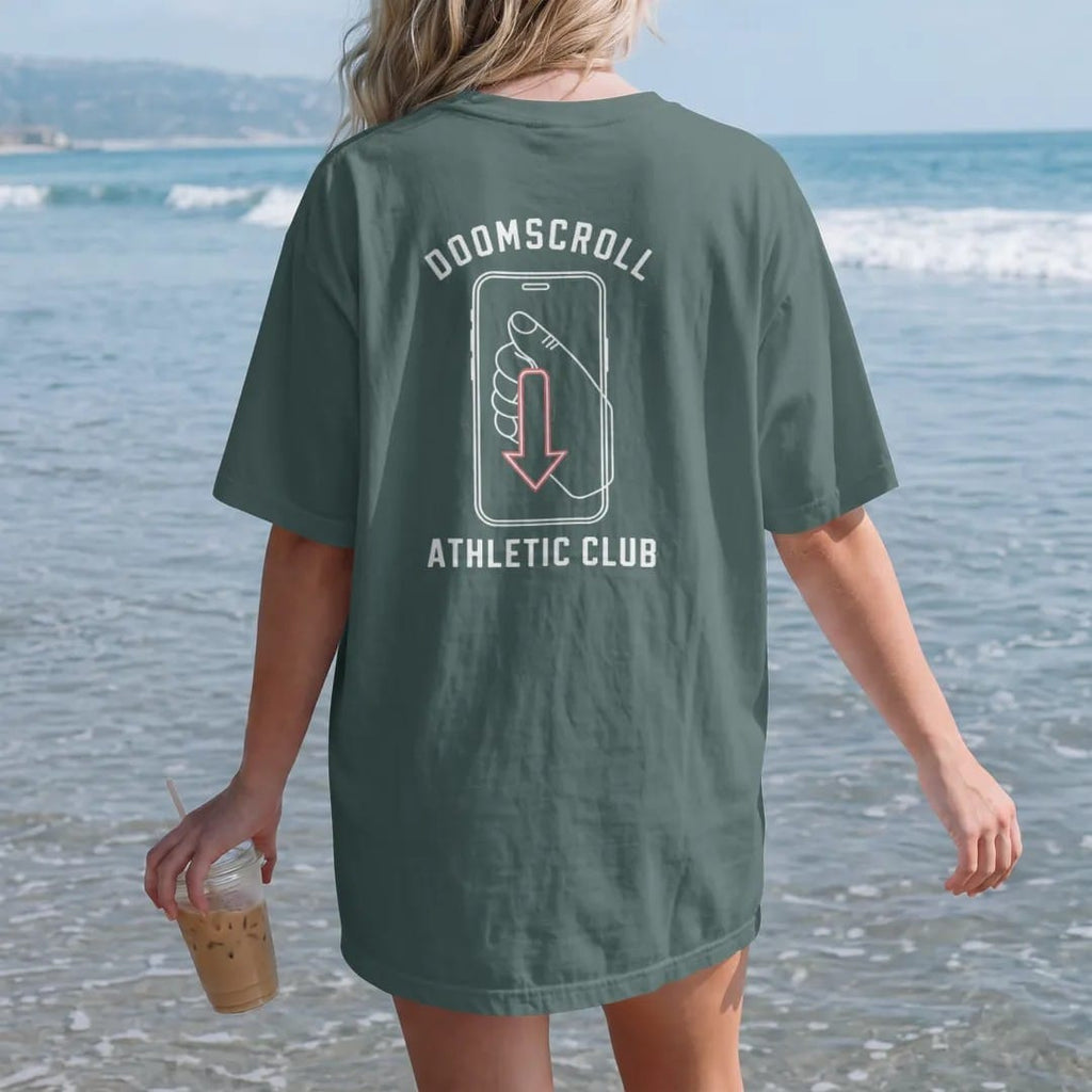 Woman on the beach in a Blue Spruce tee, back view: minimal single-line drawing of a thumb over a phone screen with a long downward scroll arrow, with "DOOMSCROLL / ATHLETIC CLUB" in small spaced-out caps, printed in bone white with a low-battery…