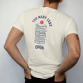 Man wearing a Ivory tee, seen from the back: minimal single-line drawing of a tall wobbly stack of browser window outlines with a loading circle on top, with "TOO MANY TABS / OPEN" in small spaced-out caps, printed in ink navy with a battery red…