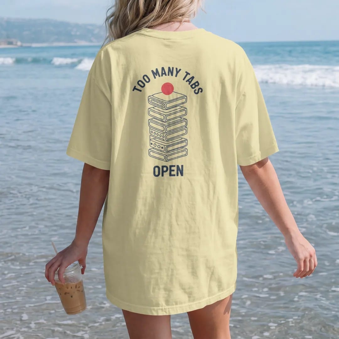 Woman on the beach in a Butter tee, back view: minimal single-line drawing of a tall wobbly stack of browser window outlines with a loading circle on top, with "TOO MANY TABS / OPEN" in small spaced-out caps, printed in ink navy with a battery red…