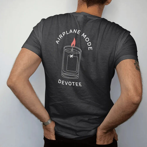 Man wearing a Black tee, seen from the back: minimal single-line drawing of a tall candle outline with a small airplane icon on the glass and a thin halo, with "AIRPLANE MODE / DEVOTEE" in small spaced-out caps, printed in bone white with a…