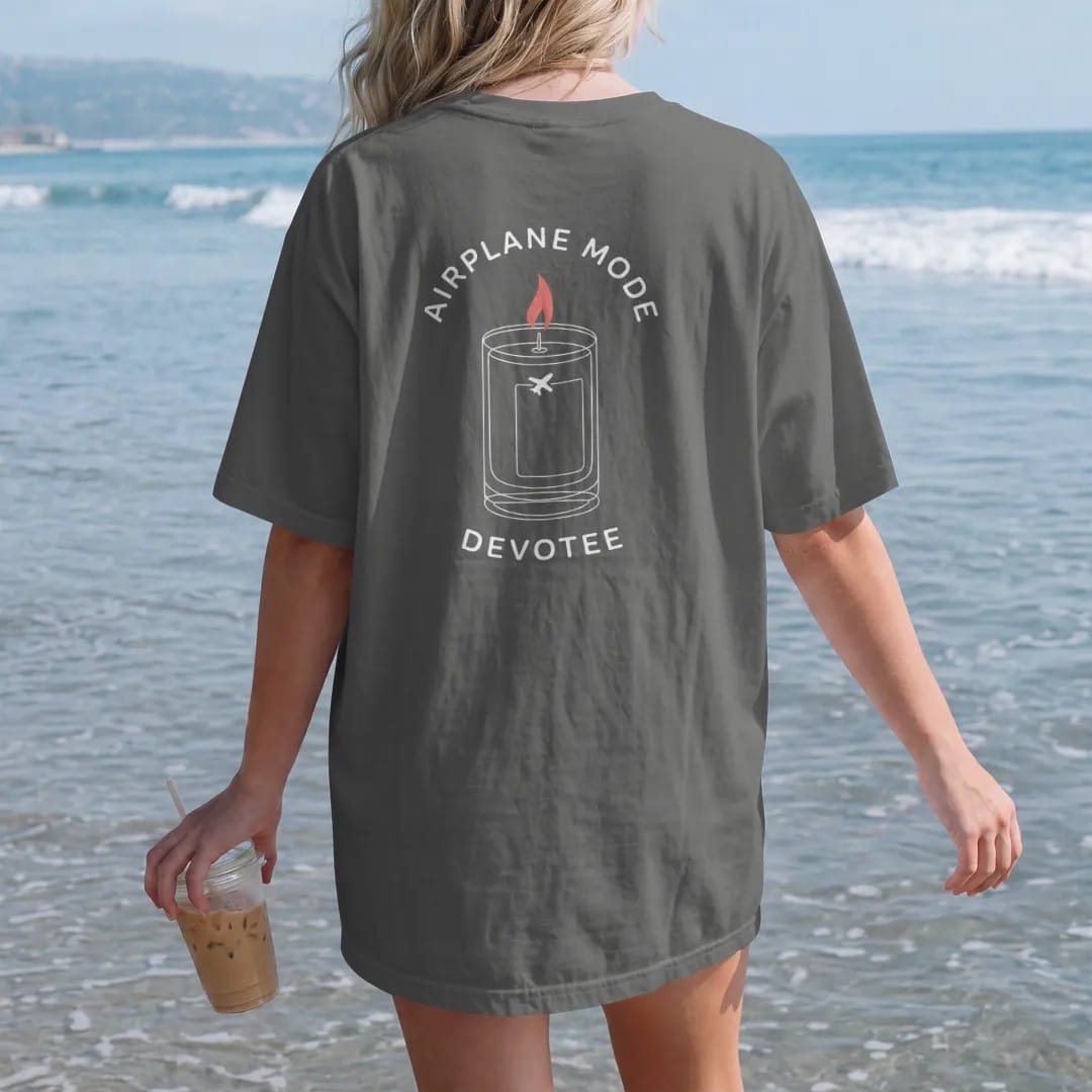 Woman on the beach in a Pepper tee, back view: minimal single-line drawing of a tall candle outline with a small airplane icon on the glass and a thin halo, with "AIRPLANE MODE / DEVOTEE" in small spaced-out caps, printed in bone white with a…