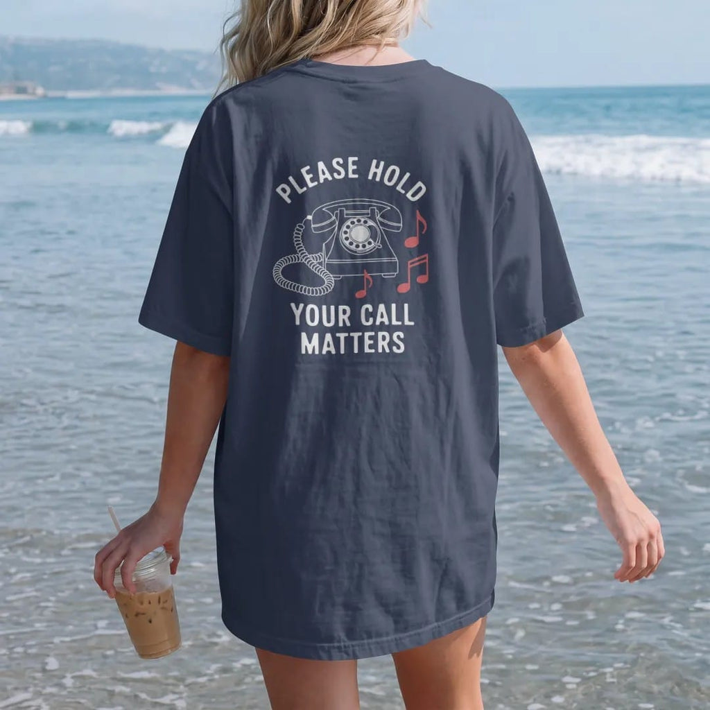 Woman on the beach in a Navy tee, back view: minimal single-line drawing of a retro desk phone whose coiled cord trails off into a few music notes, with "PLEASE HOLD / YOUR CALL MATTERS" in small spaced-out caps, printed in bone white with a…