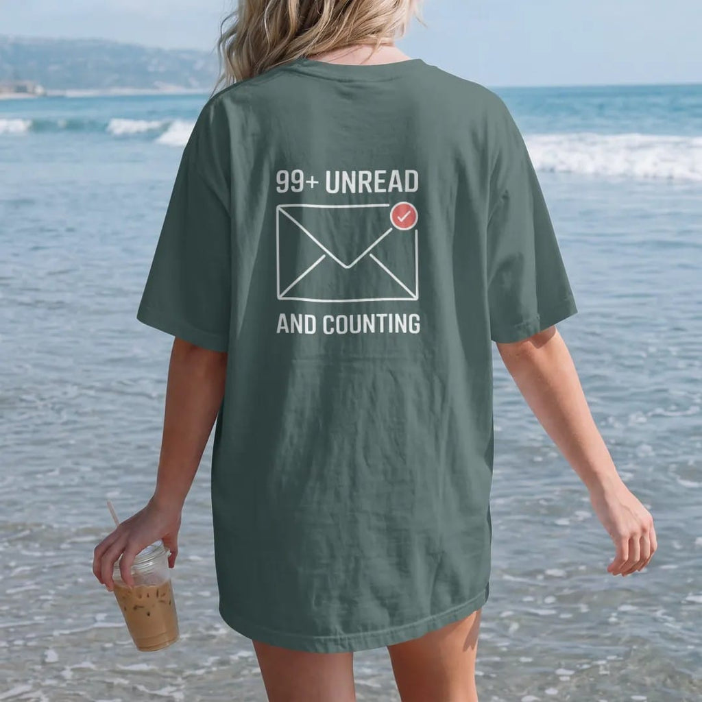 Woman on the beach in a Blue Spruce tee, back view: minimal single-line drawing of an envelope outline with a small round notification badge on its corner, with "99+ UNREAD / AND COUNTING" in small spaced-out caps, printed in bone white with a…