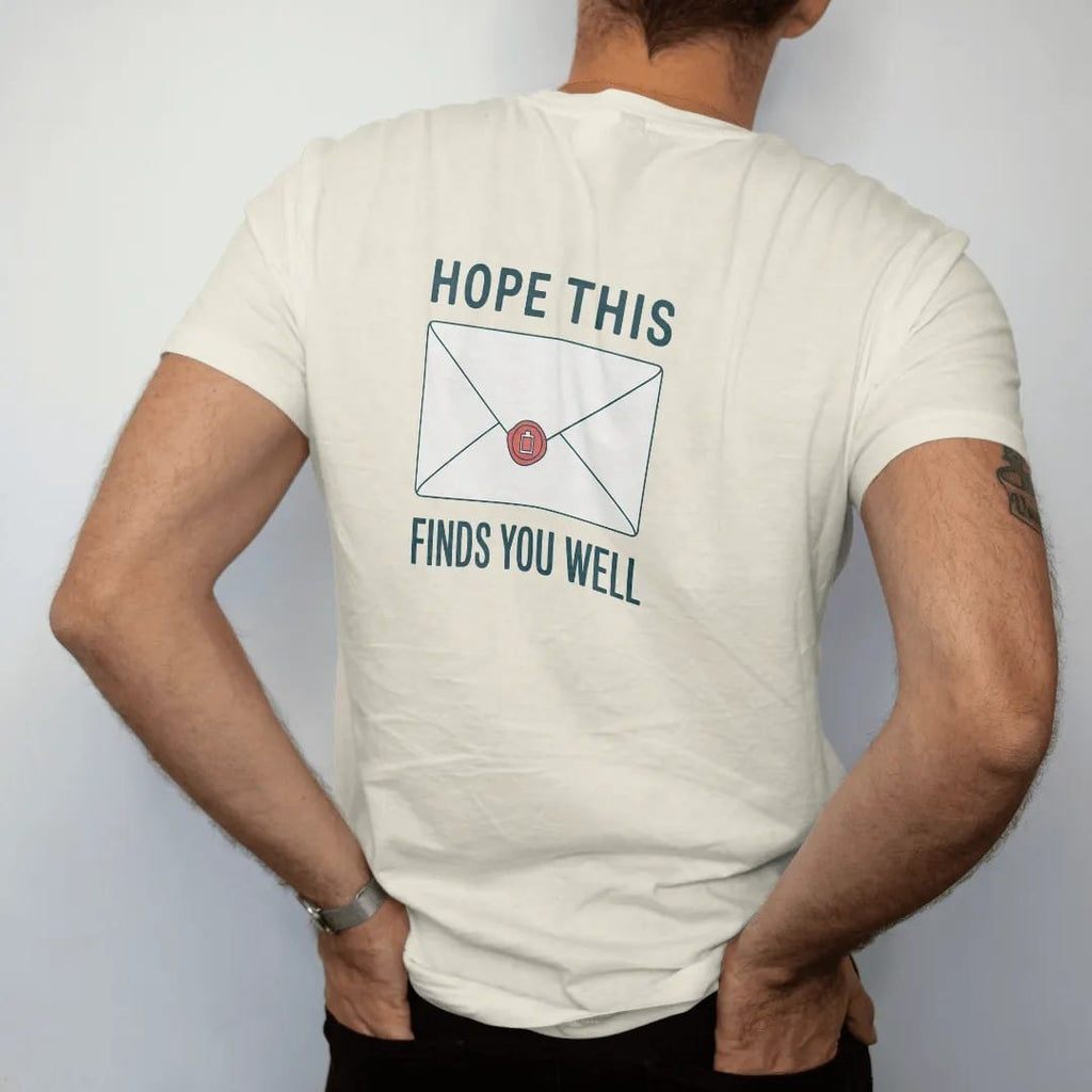Man wearing a Ivory tee, seen from the back: minimal single-line drawing of a sealed envelope with a small wax seal shaped like a battery, with "HOPE THIS / FINDS YOU WELL" in small spaced-out caps, printed in ink navy with a battery red accent on…