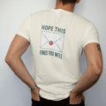 Man wearing a Ivory tee, seen from the back: minimal single-line drawing of a sealed envelope with a small wax seal shaped like a battery, with "HOPE THIS / FINDS YOU WELL" in small spaced-out caps, printed in ink navy with a battery red accent on…