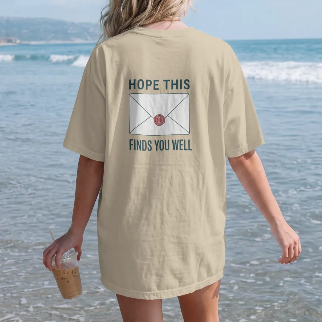 Woman on the beach in a Sandstone tee, back view: minimal single-line drawing of a sealed envelope with a small wax seal shaped like a battery, with "HOPE THIS / FINDS YOU WELL" in small spaced-out caps, printed in ink navy with a battery red…