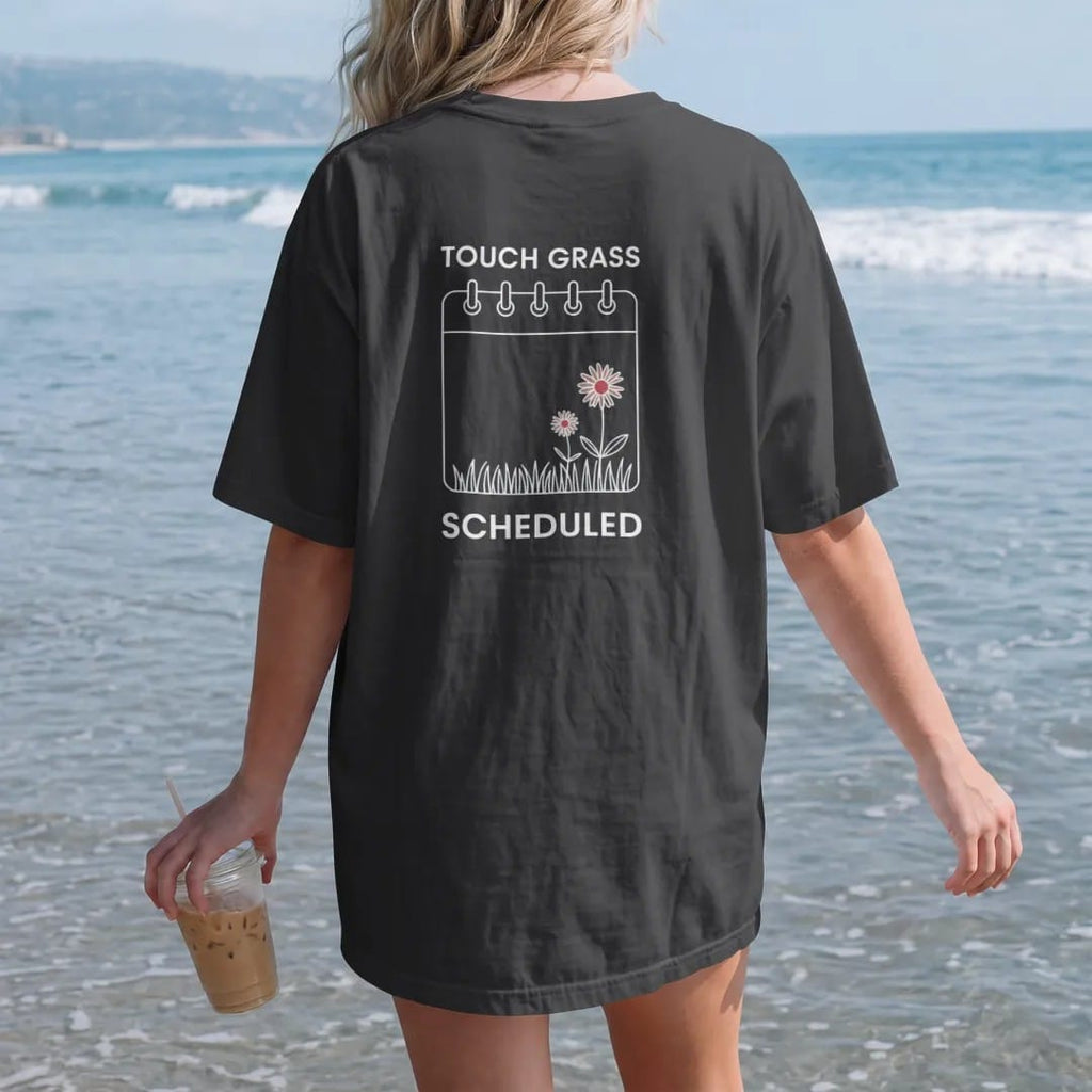 Woman on the beach in a Black tee, back view: minimal single-line drawing of a calendar invite card outline with a small tuft of grass and one daisy growing from it, with "TOUCH GRASS / SCHEDULED" in small spaced-out caps, printed in bone white…