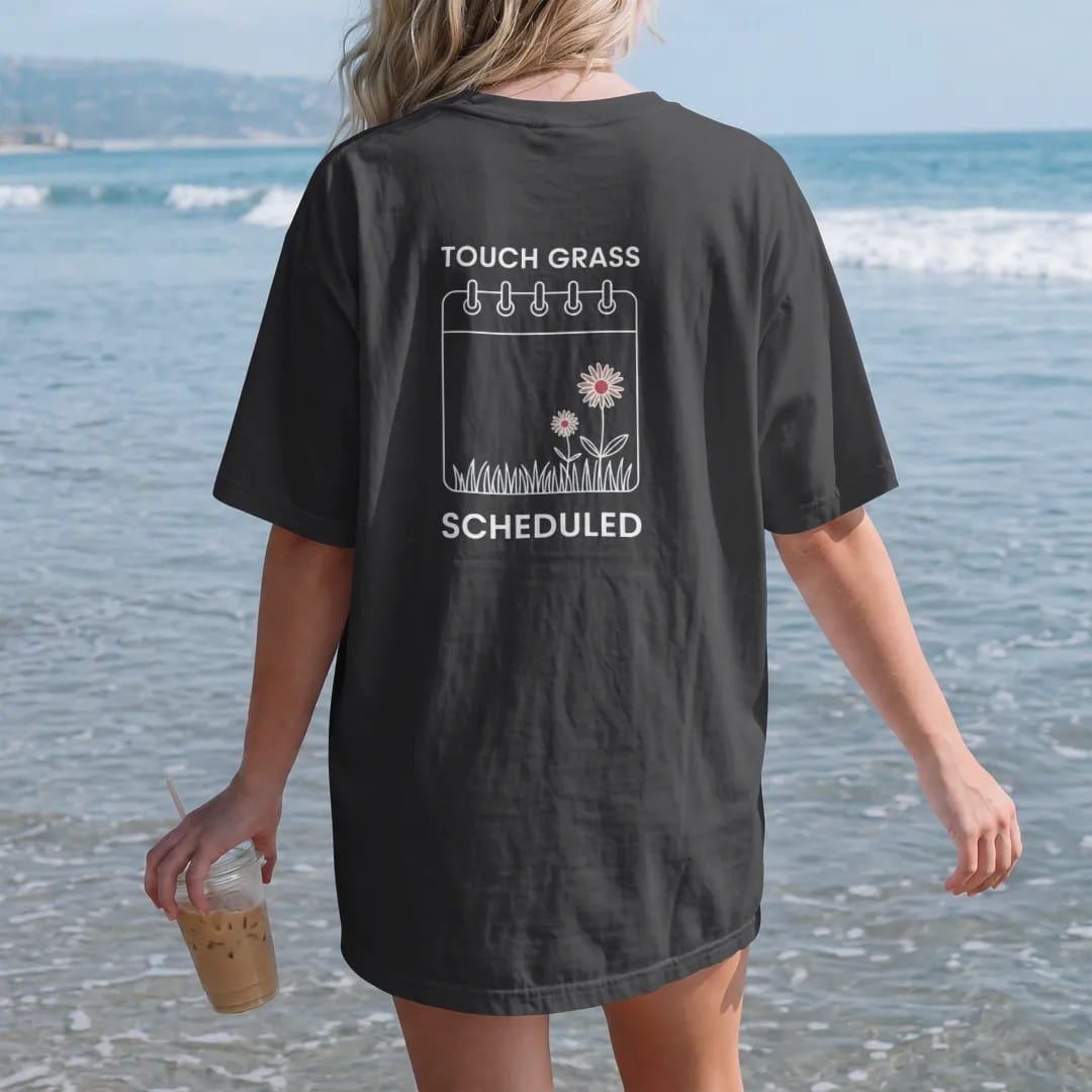 Woman on the beach in a Black tee, back view: minimal single-line drawing of a calendar invite card outline with a small tuft of grass and one daisy growing from it, with "TOUCH GRASS / SCHEDULED" in small spaced-out caps, printed in bone white…