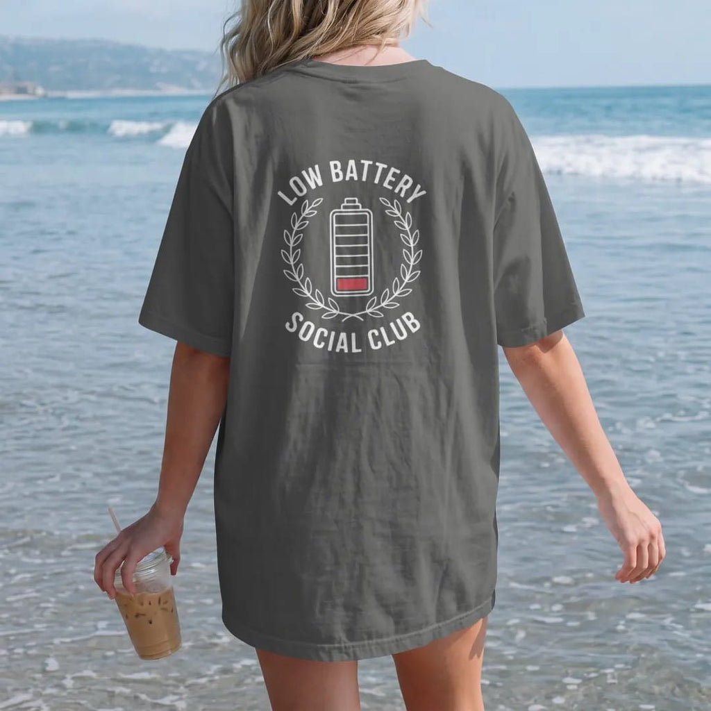 Woman on the beach in a Pepper tee, back view: minimal single-line drawing of a sleepy battery inside a thin laurel wreath, with "LOW BATTERY / SOCIAL CLUB" in small spaced-out caps, printed in bone white with a low-battery red accent on the single…