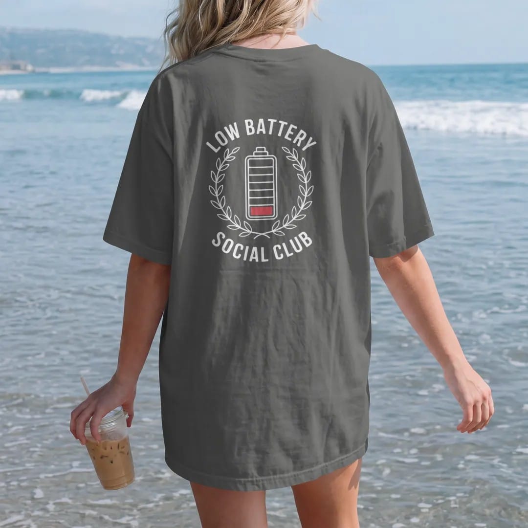 Woman on the beach in a Pepper tee, back view: minimal single-line drawing of a sleepy battery inside a thin laurel wreath, with "LOW BATTERY / SOCIAL CLUB" in small spaced-out caps, printed in bone white with a low-battery red accent on the single…