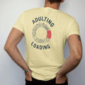 Man wearing a Butter tee, seen from the back: minimal single-line drawing of a circular loading ring made of tiny line icons: a bill, a mug, a laundry basket and a car key, with "ADULTING / LOADING" in small spaced-out caps, printed in ink navy…