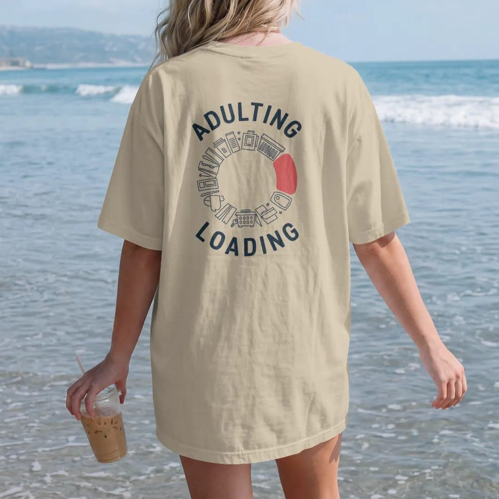 Woman on the beach in a Sandstone tee, back view: minimal single-line drawing of a circular loading ring made of tiny line icons: a bill, a mug, a laundry basket and a car key, with "ADULTING / LOADING" in small spaced-out caps, printed in ink navy…
